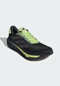 Zapatilla deportiva negra con acentos en verde neón. Presenta una parte superior de malla transpirable, superficie texturizada, diseño de tres franjas y suela acolchada.