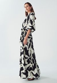 Maxi haljina u crnoj i bijeloj boji s apstraktnim uzorkom lišća, kratkim rukavima, vezanim strukom i slojevitom suknjom. Tkanina ima mekan i teksturiran izgled.