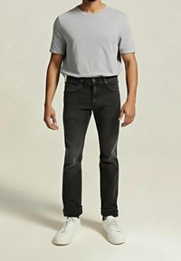 Homme portant une chemise à manches courtes gris clair, un jean noir aux revers relevés et des baskets blanches, debout devant un fond beige.
