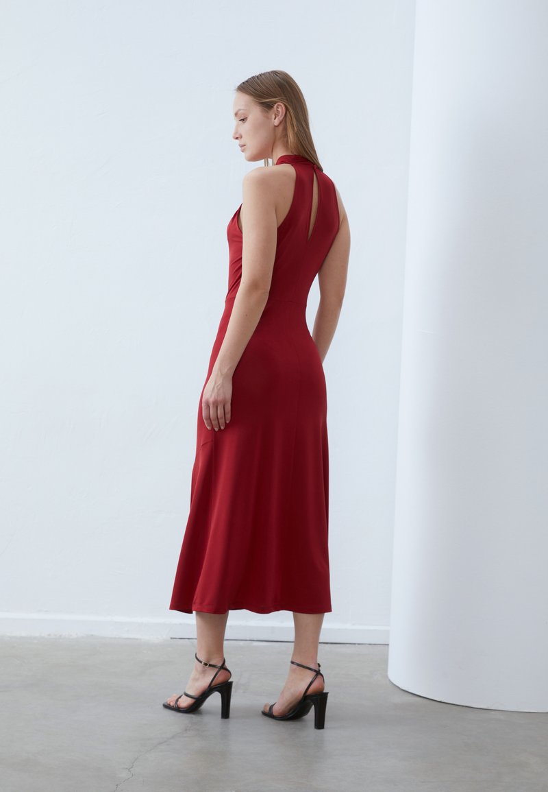 Rotes ärmelloses Kleid mit einem taillierten Oberteil, A-Linien-Rock und einem Schlüsselloch-Detail im Rücken. Getragen mit schwarzen Riemchen-High Heels. Glatte Textur.