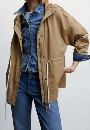 Person wearing layered clothing: blue denim jacket under tan hooded zip-up jacket, paired with blue jeans, standing with one hand behind the back.