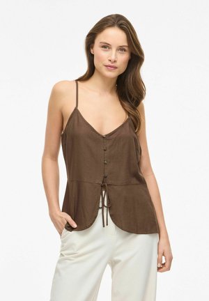 Donna con top marrone senza maniche con bottoni e bretelle sottili e pantaloni bianchi, che posa con una mano in tasca su sfondo bianco.