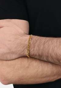 Pulsera de cadena dorada con eslabones entrelazados, que presenta una superficie pulida y una textura suave, llevada en un antebrazo sobre un fondo negro.