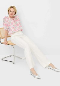 Bloemenpatroon in roze en blauw blouse, witte op maat gemaakte broek en metallic zilveren platte schoenen. Model zittend op een chroom stoel.