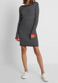 Geripptes Kleid mit schwarzen und weißen horizontalen Streifen, einem Bootsausschnitt und roten gerippten Akzenten an den Bündchen. Das Modell trägt weiße Slip-on-Schuhe.