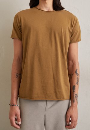 Person iført en brun kortærmet T-shirt og lysegrå bukser med synlige tatoveringer på begge underarme og et flettet armbånd.