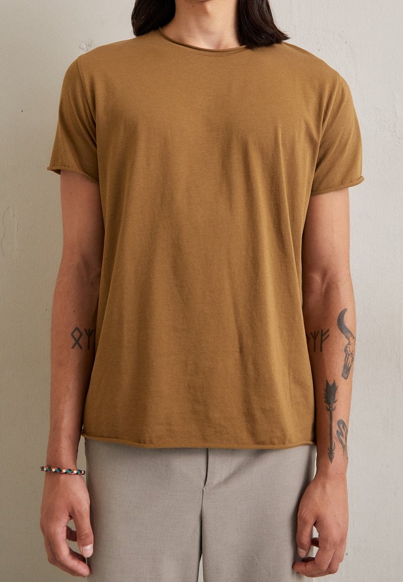 Personne portant un t-shirt marron à manches courtes et un pantalon gris clair, avec des tatouages visibles sur les deux avant-bras et un bracelet tressé.