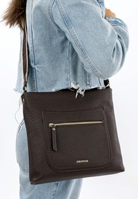 Vrouw draagt een lichtblauwe spijkerjack en jeans, en heeft een donkerbruine leren crossbodytas met textuur, voorzak met ritssluiting en logo.