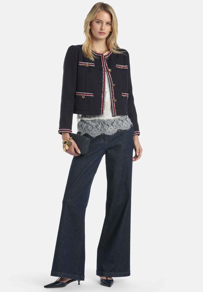 Veste marine avec boutons dorés et garniture rouge et blanche, portée sur un haut en dentelle, associée à un jean évasé foncé et des escarpins noirs à bout pointu.