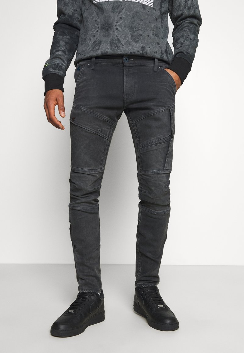 Man med svarta cargo-stil slim-fit jeans och svarta sneakers, hand i fickan, stående mot en enfärgad ljus bakgrund.