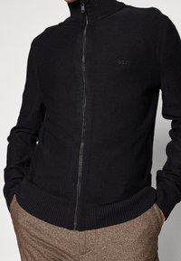 Svart zip-up stickad tröja med hög krage, ribbade manschetter och fåll; har en subtil "BOSS"-logotyp på bröstet.