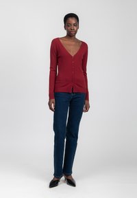 Cardigan lungo bordeaux con scollatura a V profonda e chiusura con bottoni, abbinato a jeans dritti blu scuro e tacchi neri a punta.