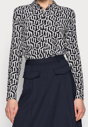 Kobieta nosząca czarno-białą, geometrycznie wzorzystą koszulę zapinaną na guziki, wpuszczoną w granatową spódnicę z wysokim stanem i kieszeniami z klapami.