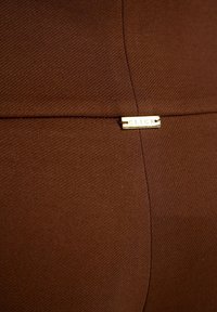 Brązowy materiał z widocznymi szwami i małą prostokątną złotą metką z wygrawerowanym słowem "CLICK", przymocowaną w miejscu przecięcia szwów.