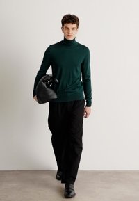 Grüner Rollkragenpullover, schmal geschnitten, leichtes Strickmaterial. Getragen mit schwarzen Hosen und einer schwarzen Tasche, vor einem neutralen Hintergrund.