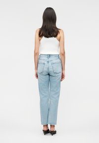 Mujer con cabello largo y oscuro, que lleva una camiseta sin mangas blanca, vaqueros de tiro alto de color azul claro y sandalias negras con tacón, de pie de espaldas.