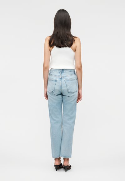 Γυναίκα με μακριά σκούρα μαλλιά που φοράει λευκό αμάνικο τοπ, ανοιχτό μπλε ψηλόμεσο τζιν και μαύρα σανδάλια με τακούνι, στέκεται με την πλάτη προς το μέρος μας.