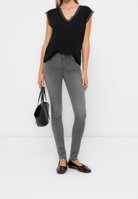 Zwarte top met kanten rand, grijze slim-fit jeans, bordeauxrode ballerina's en een zwarte handtas. De outfit contrasteert texturen en kleuren.