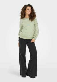 Pull vert clair avec un petit motif de cœurs dorés, col rond et ourlet côtelé, associé à un jean noir ajusté taille haute évasé et des bottes.