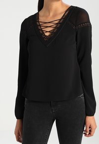 Zwarte blouse met lange mouwen en een V-hals, voorzien van een lace-up detail en doorschijnende mouwen. Glad materiaal met decoratieve afwerking bij de halslijn.