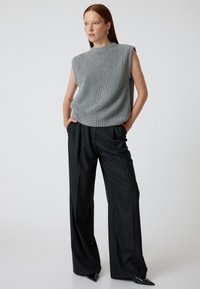Top senza maniche grigio a maglia con un motivo testurizzato, abbinato a pantaloni larghi neri a vita alta. Il modello si presenta con sicurezza davanti a uno sfondo semplice.