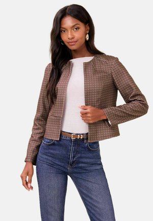 Woman wearing a brown checkered cropped jacket, white top, blue jeans, and gold hoop earrings against a plain white background.