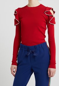 Maglietta rossa a maniche lunghe con spalle a strisce e bordo bianco. Abbinata a pantaloni blu con cordoncino e strisce laterali di colori diversi.