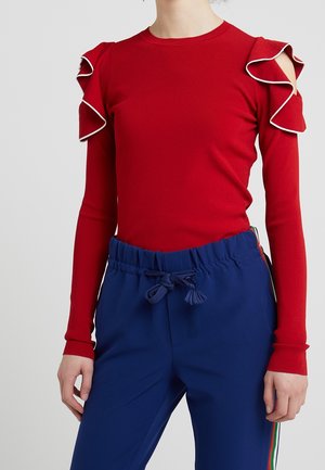 Maglietta rossa a maniche lunghe con spalle a strisce e bordo bianco. Abbinata a pantaloni blu con cordoncino e strisce laterali di colori diversi.