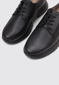 Zapatos de cuero negro con cordones, suelas texturizadas y punteras redondeadas, exhibidos sobre un fondo gris.