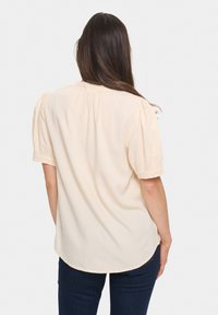 Blusa color crema con maniche corte a sbuffo, tessuto testurizzato e orlo arrotondato. Presenta un collo arricciato e un motivo sottile.