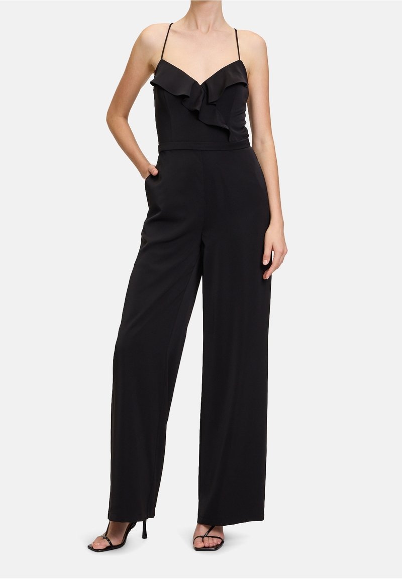 Femme portant une combinaison noire sans manches avec un corsage à col en V volanté, un pantalon à jambes larges et des sandales noires à talons hauts, debout avec une main dans la poche.