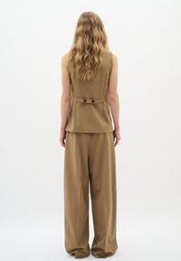 Gilet su misura color tan con dettagli a bottoni sul retro e pantaloni a gamba larga abbinati. Tessuto liscio e strutturato con un design formale.