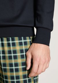 Chemise noire à manches longues avec des poignets côtelés, associée à un pantalon à carreaux comportant des motifs à carreaux verts, noirs, jaunes et blancs. Main visible.