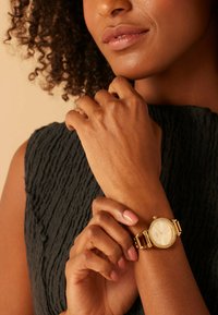 Frau mit lockigem Haar, die ein strukturiertes schwarzes Oberteil trägt und eine goldene Armbanduhr mit rundem Zifferblatt und Kristallfassung an ihrem linken Handgelenk justiert.