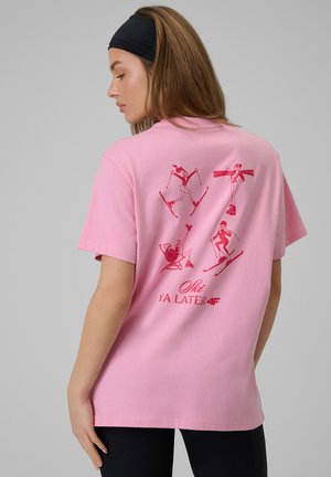Kobieta w różowej koszulce z czerwonymi ilustracjami tematyki narciarskiej i napisem „Ski YA LATER” na plecach, w połączeniu z czarnymi legginsami i czarną opaską na głowę.