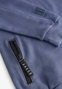 Modrá mikina s hladkým povrchom, obsahuje zips, ktorý má čierne kovanie, a etiketu s nápisom "BAKER" a dizajnom čiarového kódu.