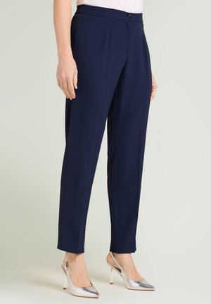 Mujer con pantalones a medida azul marino hasta el tobillo y zapatos de tacón plateados con tira trasera, de pie contra un fondo liso.