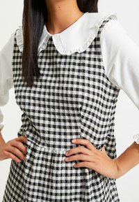 Svart och vit rutig klänning med en rynkad vit krage och åtsittande rabatt. Tyget har en strukturerad yta och är åtsnörpt i midjan.