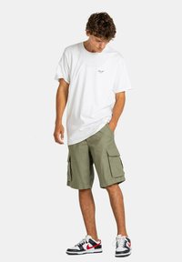 Camiseta blanca de algodón, de corte holgado, con un pequeño detalle de logo. Pantalones cortos cargo verde oliva con bolsillos laterales. Zapatillas negras y rojas con detalles blancos.