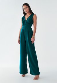 Teal jumpsuit med dyb V-udskæring, brede ben og en tætsiddende talje. Lavet af glat stof, har et minimalistisk design uden mønstre.