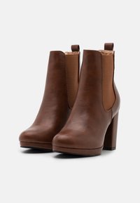 Bottines en cuir marron avec un talon épais, des panneaux latéraux élastiques et une texture lisse. Présentent un bout rond et une languette de tirage.