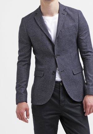 Mand iført en tætsiddende mørkegrå blazer over en hvid t-shirt og sorte bukser, stående mod en ensfarvet lys baggrund.