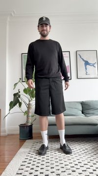 Black long-sleeve shirt, black shorts, white socks, and black shoes. Indoor setting with a plant and sofa in the background.