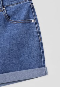 Shorts en denim bleu avec ourlet retroussé et poche avant, montrant des coutures détaillées et un rivet métallique à l'angle de la poche.