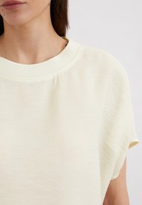 Vaaleankeltainen, lyhythihainen paita, joka on valmistettu tekstuuripinnasta. Siinä on leveä pääntie ja rento leikkuu, ja siinä on hillitty ribbineulostammi.