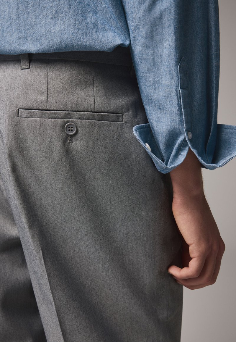 Pantaloni grigi con una texture liscia presentano una tasca posteriore con bottone. Una camicia blu con una texture presenta polsini arrotolati.