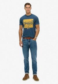 Blauw katoenen T-shirt met geel grafisch logo, gecombineerd met lichtblauwe jeans en bruine laarzen. Korte mouwen en een ronde hals.