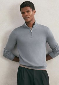 Mężczyzna w jasnoszarym swetrze z krótkim suwakiem na kołnierzu, nałożonym na beżową koszulę i czarne spodnie, stoi na tle neutralnej, teksturowanej ściany.