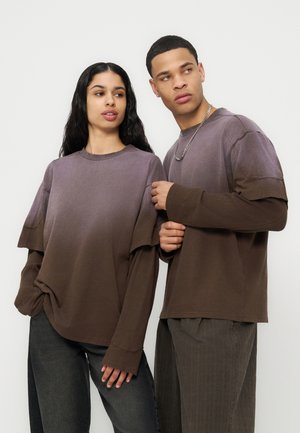 Jonge man en vrouw dragen bruine gradient lange mouw tops in laagjes en donkere broek, staand tegen een effen lichte achtergrond.