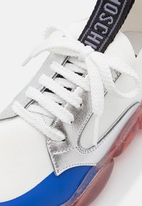 Sko med en blandning av vitt textilmaterial, metalliska silverdetaljer, blå sektioner och en genomskinlig röd sula. Inkluderar vita skosnören och ett svart spänne.